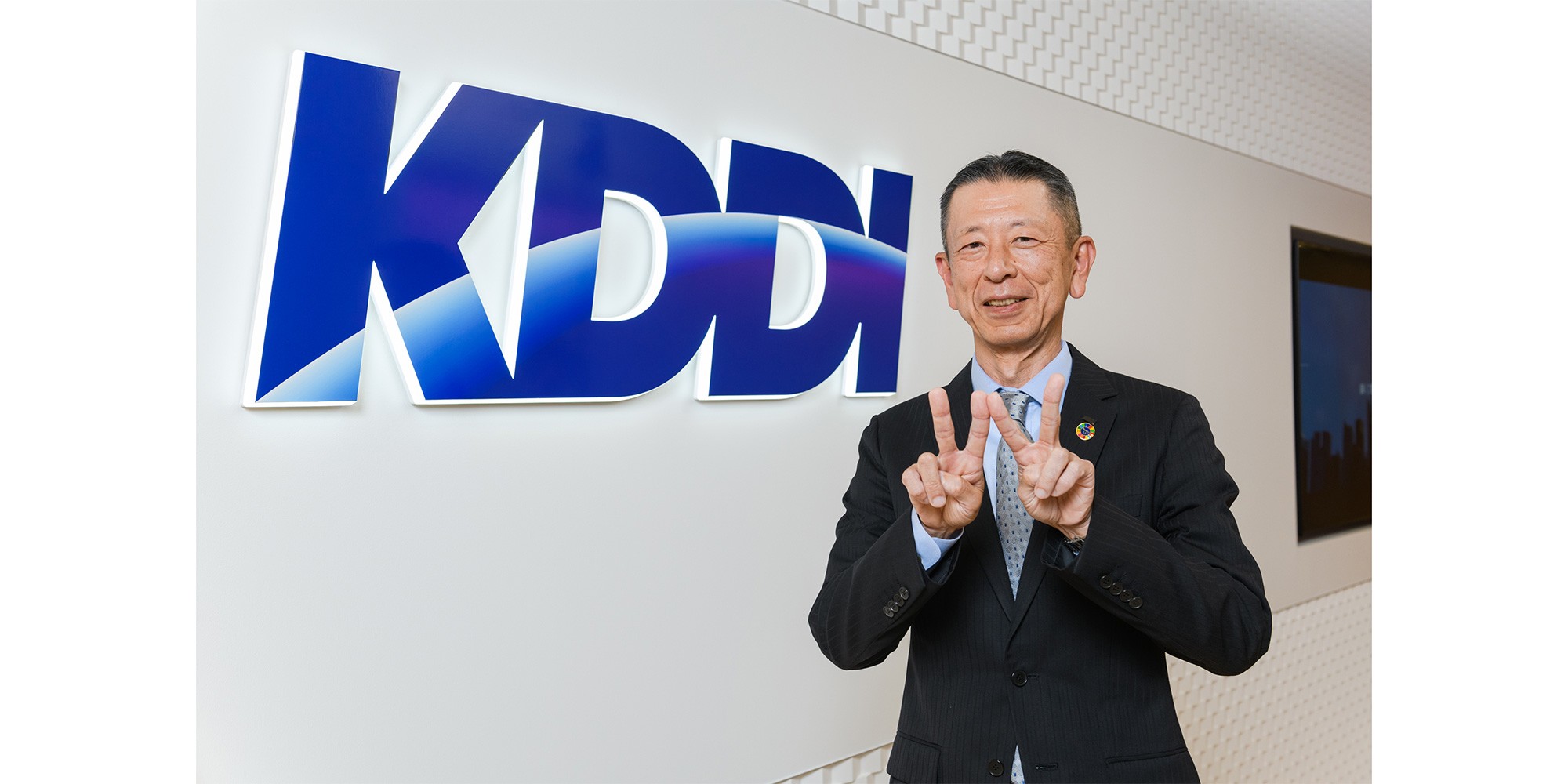 KDDIロゴの前で桑原副社長が「W」サインをしている写真