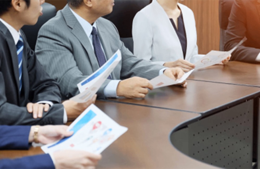 議事録ツールの利用シーンを示す画像。経営会議や管理職会議で複数のビジネスマンが会議テーブルを囲み、資料や議事録を確認しながら議論している様子。