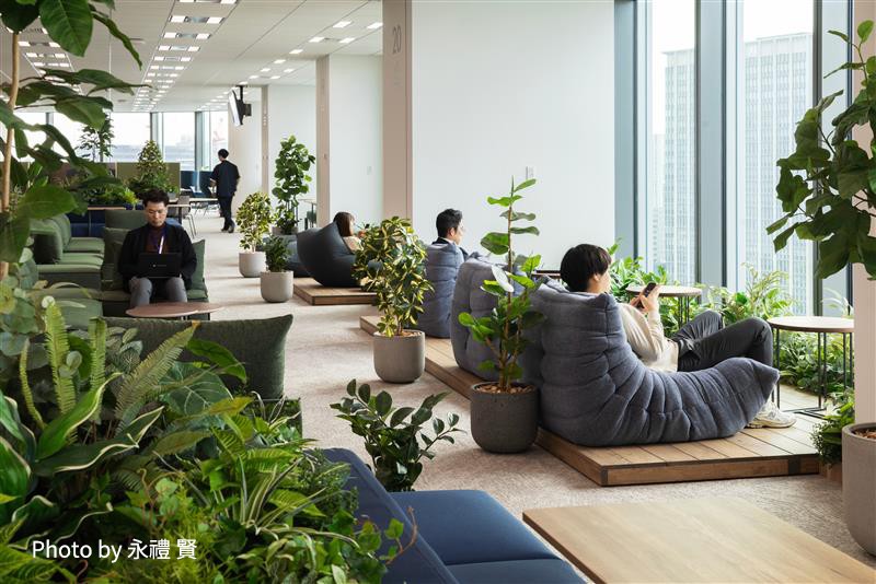 大きな窓から自然光が差し込む開放的なラウンジスペースで、数人の人がリラックスしながらくつろいでいる様子。多くの観葉植物が配置されている。（Photo by 永禮 賢）