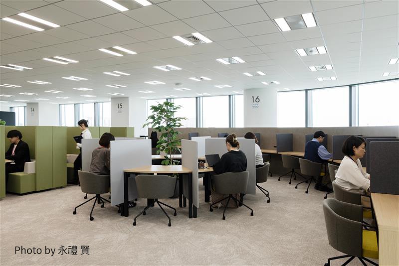 多くの人がパーテーションや仕切りで区切られたオフィスやコワーキングスペースで作業している様子。大きな窓から自然光が入る明るい環境。（Photo by 永禮 賢）
