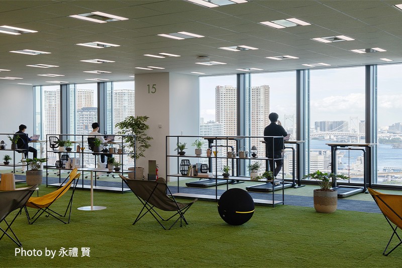 芝生エリアにバランスボールや植物が配置されており、その奥にランニングマシンやサイクリングマシンを使いながら仕事をしている人々の様子。リラックスしながら運動もできるカジュアルなオフィス環境。（Photo by 永禮 賢）
