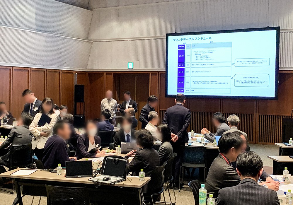 会議室で多くの参加者が座っている様子。スクリーンにはタイムテーブルのスケジュールが表示されている。参加者同士が議論や資料の確認をしている場面。
